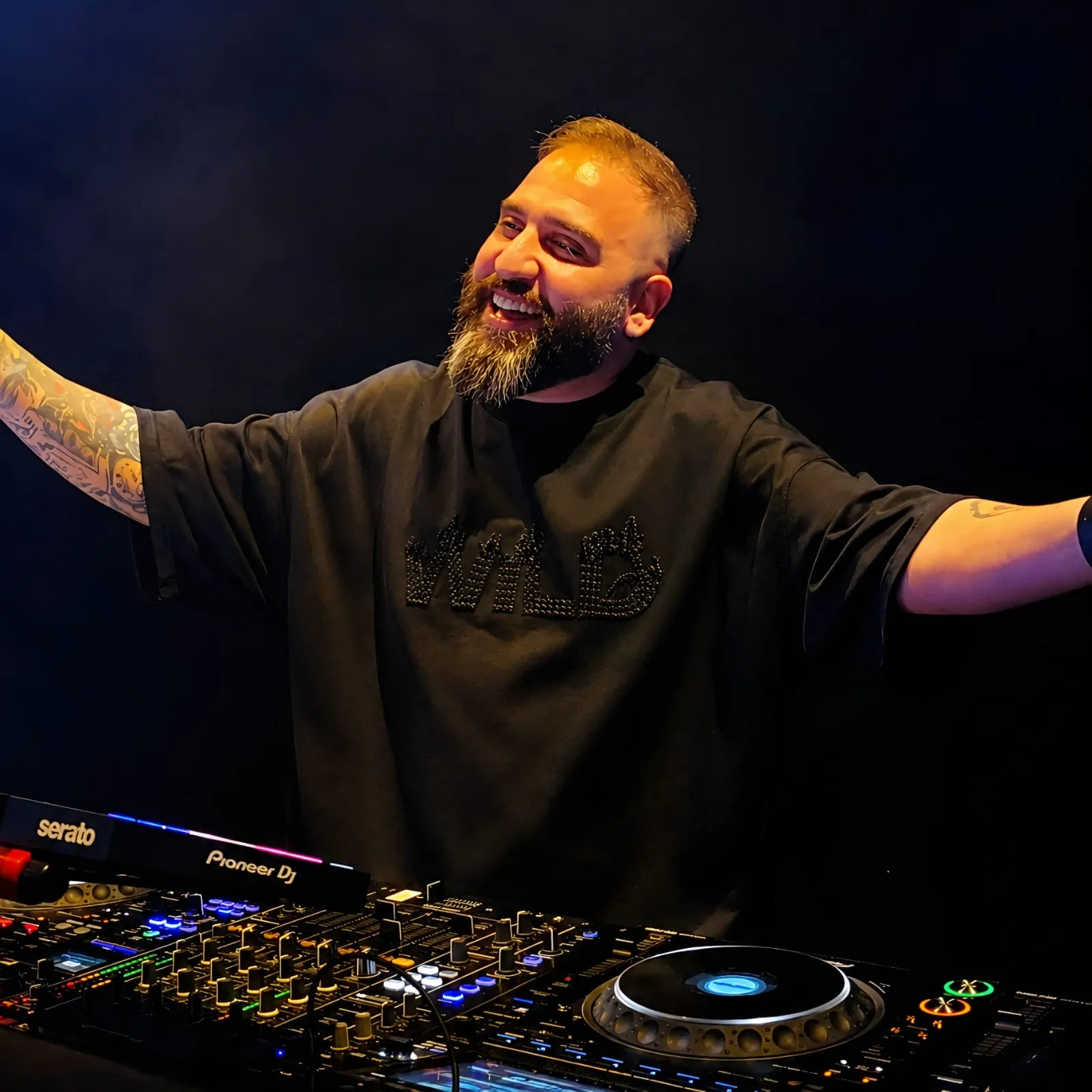 Bearded DJ in a black tee with both arms spread wide behind the decks