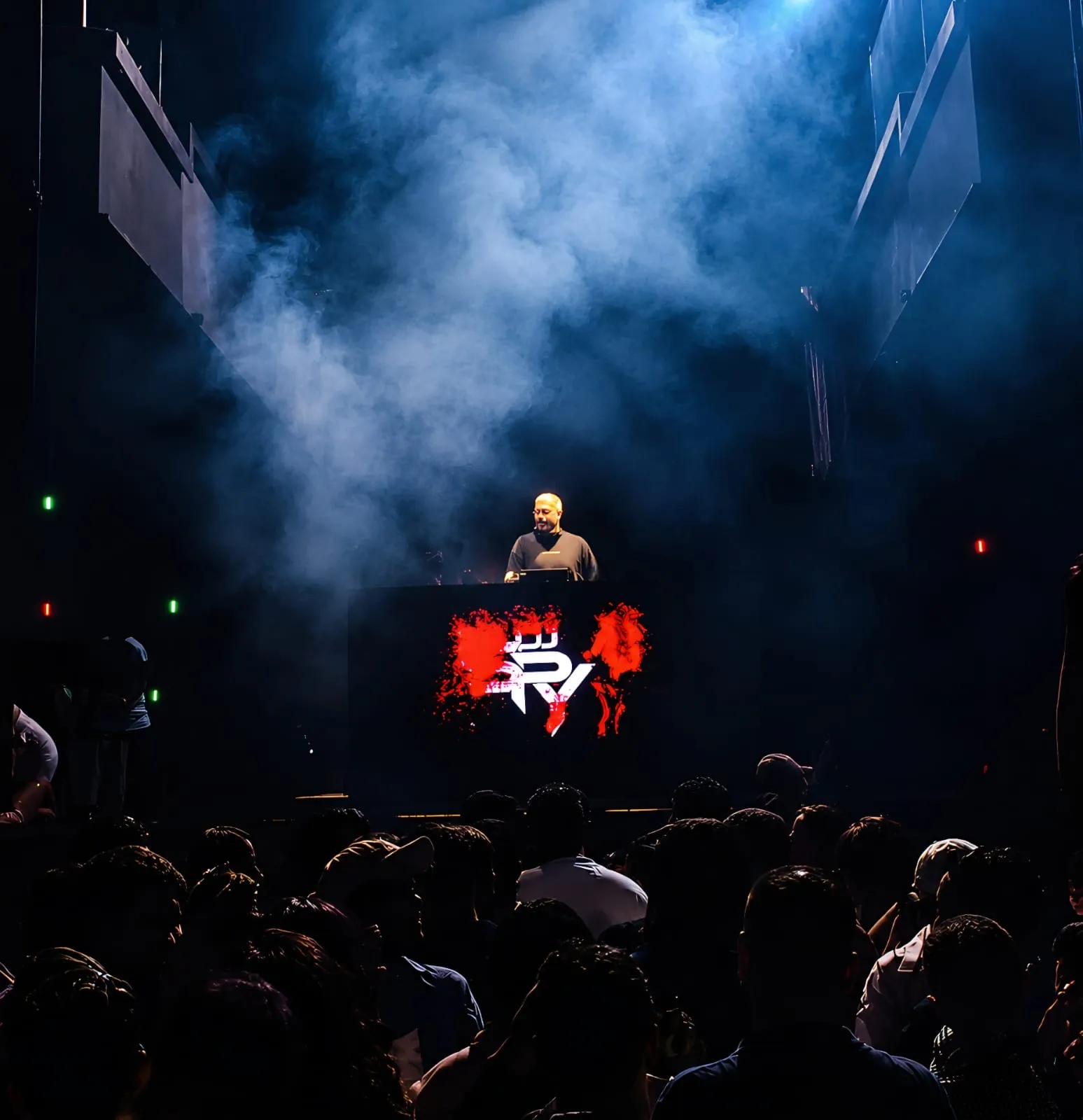DJ booth glowing red in the dark as a crowd faces the stage through blue haze
