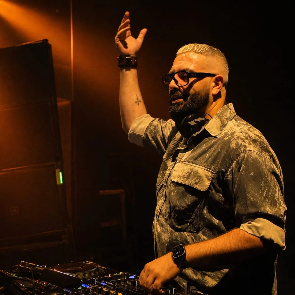 DJ in a camouflage shirt with a hand raised beside a tall speaker in warm light