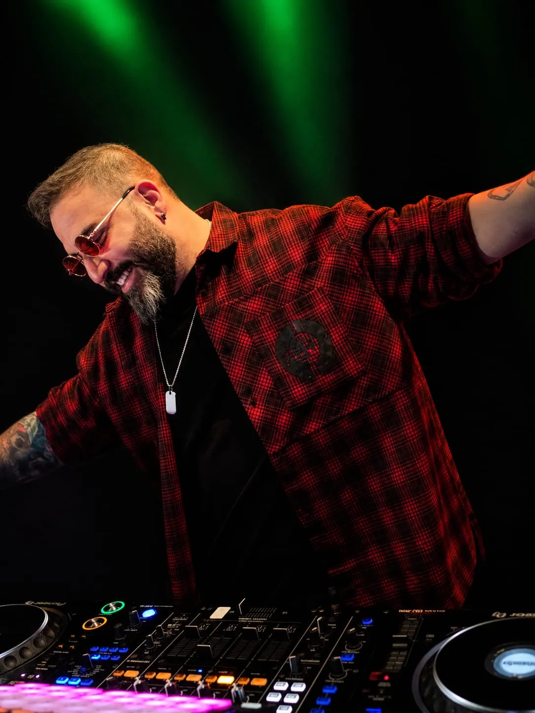 DJ in a red plaid shirt smiling with one arm out under green lights
